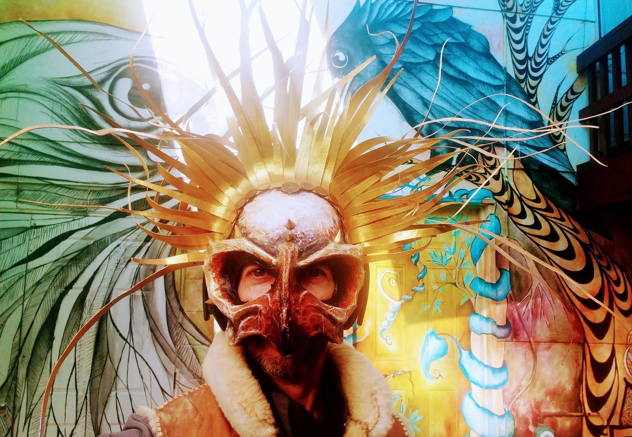 Christian Borrego wearing a gold skull mask and dramatic headdress in front of a colorful mural